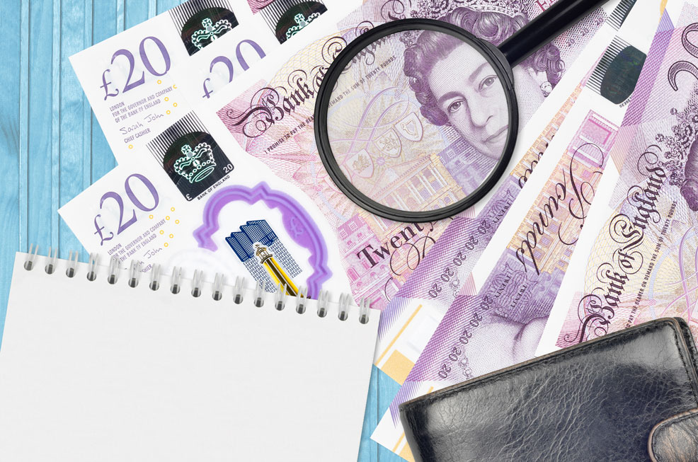 A flat lay image featuring several £20 Bank of England notes arranged on a light blue wooden surface, with a magnifying glass positioned over the banknotes. To the left is a notepad with blank white pages ready for information to be added. The overall composition suggests investigation or close examination of business expenses.